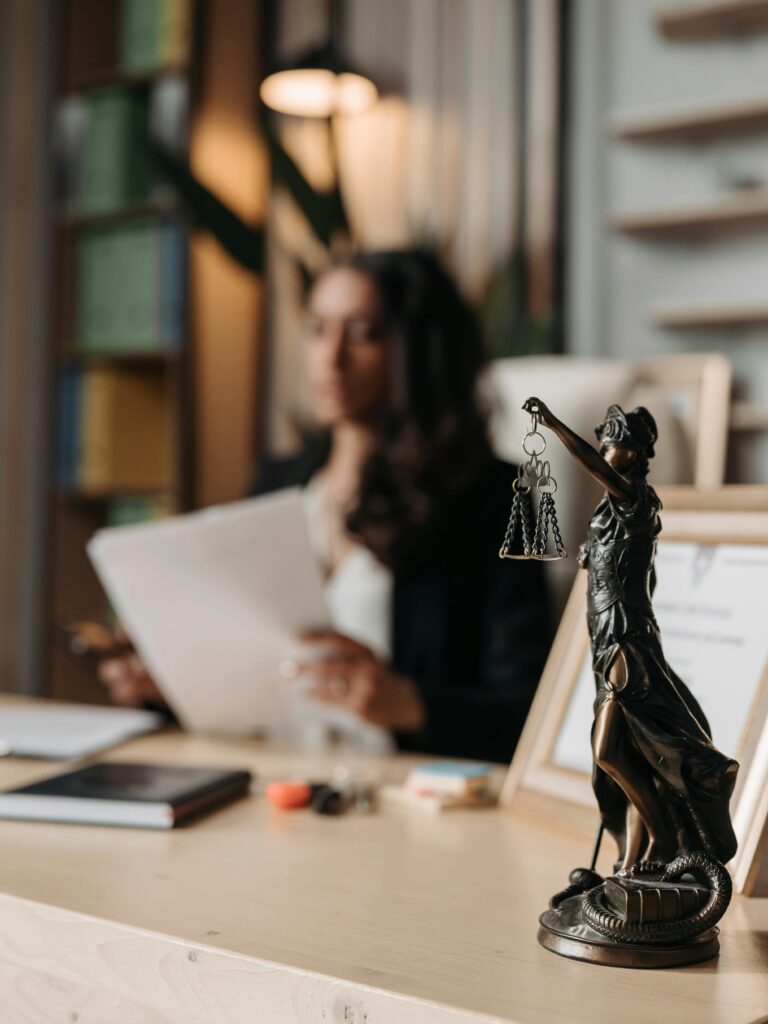 Close-up of a justice figurine on desk with blurred female lawyer in office setting.